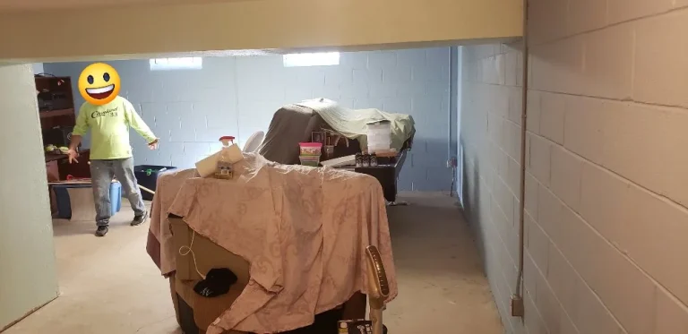 Homeowner mid-project painting basement walls, with furniture covered in sheets and one wall showing the transition from beige to blue-gray paint