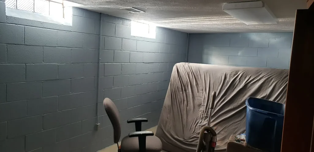Basement cinder block walls freshly painted in blue-gray with furniture covered by a drop cloth during a DIY painting project