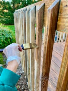 A wooden fence being coated with solid stain over old paint.