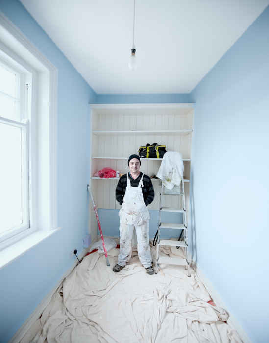 A painter standing in a freshly painted room with tools in the background.