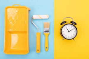 Painting tools like brushes, rollers, and tape placed beside a clock to symbolize the time cost of DIY painting.